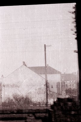 Vintage black-and-white urban street scene featuring a low-angle view of a single-story building with a flat roof and visible...