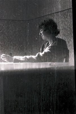 Mid-century modern interior shot: woman in a sleeveless blouse sits at a minimalist glass-topped desk, writing with a fountai...