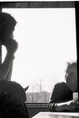 Silhouetted figure leans against a window frame, while another person in a striped sweater holds a cup, both facing outdoor l...