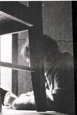 Black-and-white interior shot of a child peering through wooden blinds at an unseen outdoor scene, with a cat resting on the ...