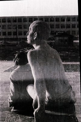 Monolithic concrete statue of seated, shirtless figure with head tilted upward, set against industrial backdrop. Likely 1960s...