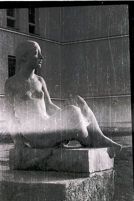 Neoclassical seated female fountain statue in monochrome, water cascading from back. Likely 20th-century urban public art, Eu...