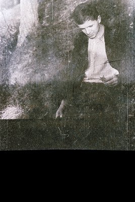 Vintage black-and-white photo of a young man in a checkered shirt, seated on a stone step or ledge, holding a small object. T...