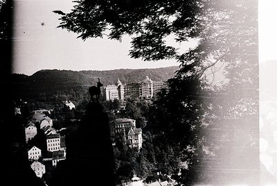 Vintage black-and-white aerial view of a European hillside town with grand, multi-story buildings and lush greenery. Prominen...