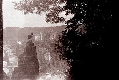Vintage sepia-toned statue of a deer atop a rocky outcrop overlooking a European cityscape, likely early 20th century. Archit...