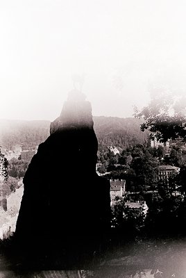 Silhouette of a person in a long coat or cloak facing a misty, forested hillside town. Architectural details include a promin...