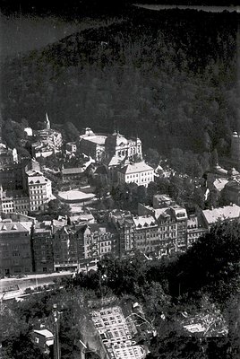 Aerial view of a historic European town nestled in a mountainous valley, likely from the mid-20th century. Prominent multi-st...