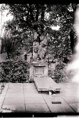 Neoclassical tomb monument featuring a draped female figure in relief, flanked by urns and inscribed plaques, set in a lush, ...