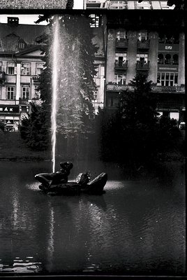 Black-and-white urban fountain scene featuring a child in a small boat beneath a powerful vertical water jet. Surrounding bui...