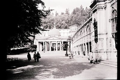 Neoclassical courtyard with ornate columns and arched walkways, likely a historic spa or resort. Three figures in vintage att...