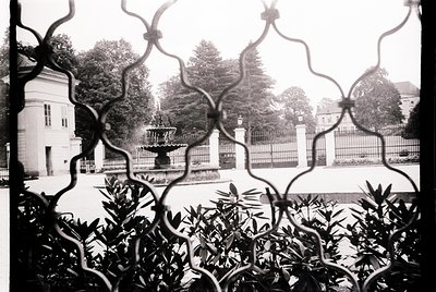 Classic black-and-white courtyard view through ornate wrought-iron lattice, showcasing a symmetrical fountain and classical a...