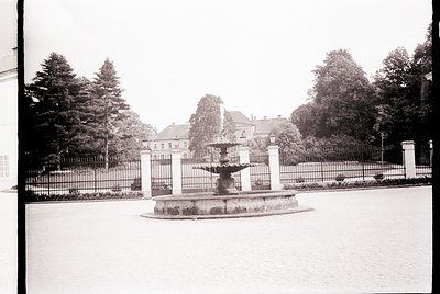 Neoclassical fountain in a landscaped courtyard, surrounded by tall coniferous trees and a wrought-iron fence. The central st...