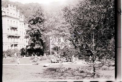Vintage black-and-white park scene featuring early 20th-century European architecture. Prominent multi-story building with or...