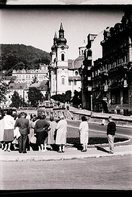 Vintage black-and-white photo of a group of mid-20th-century tourists observing a Baroque-style church with twin towers and d...