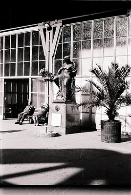Neoclassical bronze statue of a woman holding a wreath atop a pedestal in an urban courtyard, flanked by potted palm trees. T...