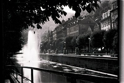 Classic black-and-white shot of a grand fountain in a European city square, likely 1950s–1960s. Water jets arch dramatically ...