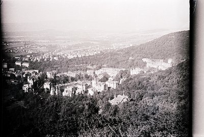 Vintage aerial view of a forested hillside town with scattered buildings and a prominent castle-like structure. Dense greener...