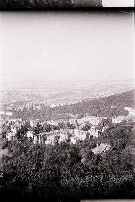 Vintage aerial view of a densely forested hillside town with medieval-style architecture, likely European. Prominent stone st...