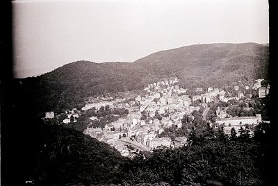 Vintage black-and-white aerial view of a hillside town nestled between forested slopes, likely early-to-mid 20th century. Den...