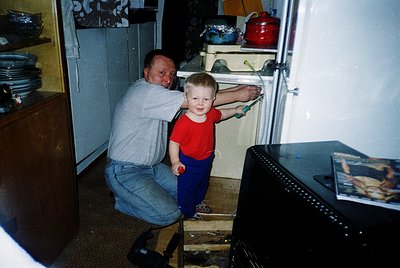 Vintage kitchen scene featuring a man kneeling beside a young boy in a red shirt and blue pants, examining an open refrigerat...