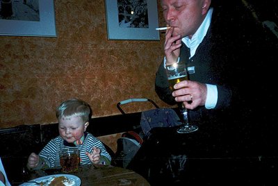 Vintage indoor dining scene: man in 1970s-style vest and tie smokes while sipping from a wineglass; toddler in striped shirt ...