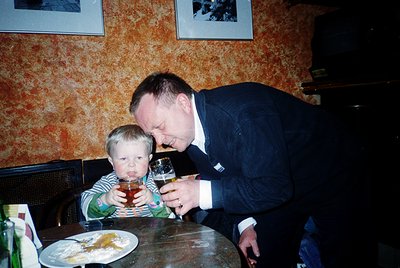 A man in formal attire leans over a wooden table, offering a wineglass to a young child seated beside him. The child, wearing...