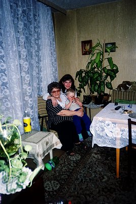 Two women in a mid-20th-century interior, likely Eastern Europe (1960s–1980s). A seated woman holds a baby, surrounded by flo...