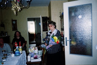 Vintage indoor celebration featuring a woman in a black-and-white patterned dress holding a floral bouquet, likely a birthday...