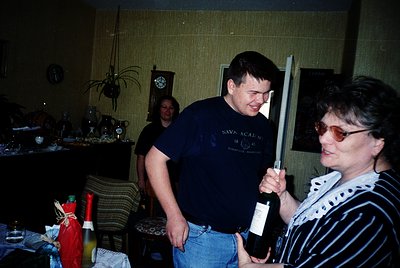 Vintage indoor gathering with two adults holding wine bottles, one wearing a 1990s-style "Navy Academy" polo shirt. Decor inc...