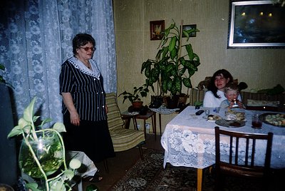Vintage indoor scene featuring two generations in a modest living/dining space, likely Eastern Europe, 1970s-1980s. Elderly w...