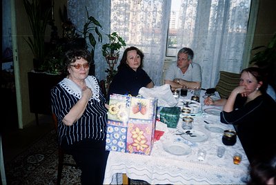 Vintage indoor gathering featuring four adults seated around a table draped in a floral-patterned tablecloth. The setting app...