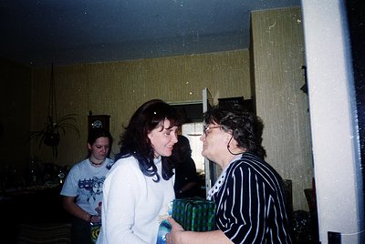 Vintage indoor gathering featuring three women in casual 1990s attire—one holding a wrapped gift. Warm lighting, floral arran...