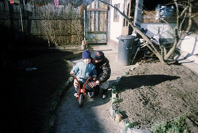 A man in a dark jacket assists a child on a red bicycle in a residential courtyard. The setting appears to be a modest urban ...