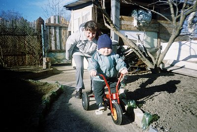 A woman assists a toddler on a red balance bike in a residential backyard, likely . Bare trees and a wooden fence suggest lat...