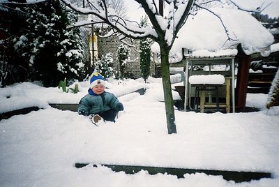 Child in winter attire sits in snow-covered garden, surrounded by snow-laden trees and traditional wooden structures. Likely