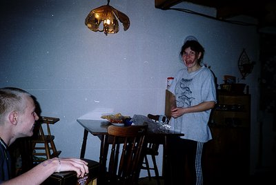Vintage indoor scene featuring two individuals in a dimly lit room with a wooden table and chairs. One person holds a Wii rem...