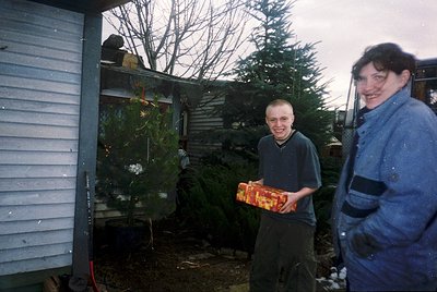 Two individuals pose outdoors near a mobile home, holding wrapped gifts. The younger person wears a dark sweater and holds a ...