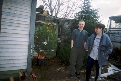 Two individuals stand outdoors near a small wooden shed and a decorated Christmas tree in a backyard setting. The tree, adorn...