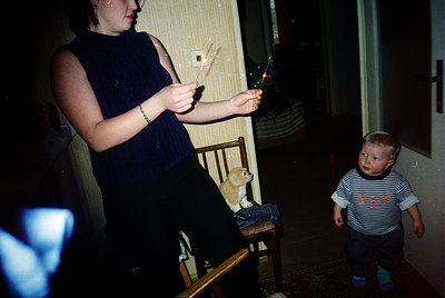 Vintage indoor scene: pregnant woman in a sleeveless navy top holds a lit sparkler, standing beside a wooden chair. A small c...