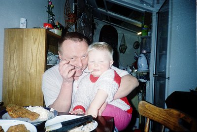 A man and child share dessert in a vintage kitchen, likely mid-1990s. The man feeds the child a spoonful of cake, while a pla...