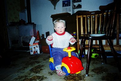 Vintage 1990s indoor shot of toddler in a red/yellow push toy car, wearing striped sweater and patterned pants. Wooden chair,...