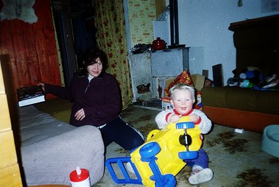 Vintage indoor portrait of a woman and child in a modest living space, likely mid-1980s–1990s. The woman sits on a worn sofa ...