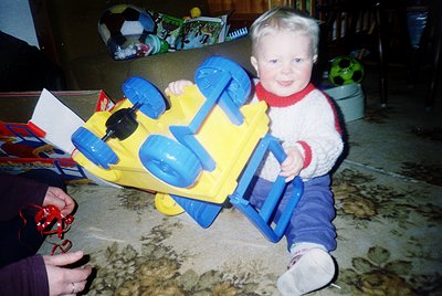 Vintage plastic toy truck with blue wheels and yellow body, held by a smiling toddler in a red-and-white sweater. Indoor sett...