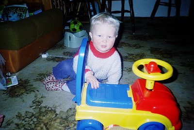 Vintage childhood scene: toddler in 1980s-style knit sweater and patterned pants plays with a bright yellow/red/blue toy truc...