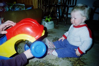Vintage plastic toy truck (red/yellow/blue) with blue wheels, pushed by a toddler in a white sweater with red trim, patterned...