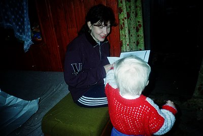 A woman in a navy sweatshirt with a "V" logo sits on a green cushion, reading to a young child in a red knit sweater. Warm, d...