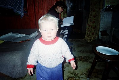 Young child in 1970s-style knit sweater and pants, standing indoors in dim lighting. Wooden floor and basic furnishings sugge...