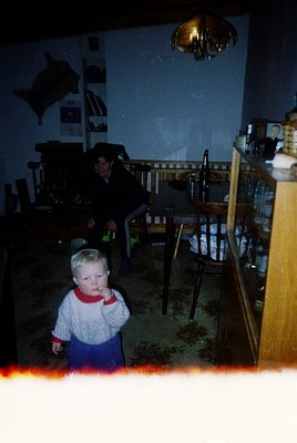 Vintage indoor scene featuring a young child in a knitted sweater and blue pants, standing near a wooden railing. Adult in da...