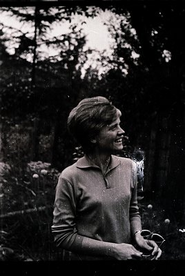 Vintage black-and-white portrait of a woman in a forest setting, mid-laugh, wearing a collared sweater and holding a small ob...