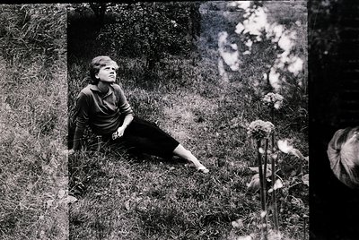 Black-and-white portrait of a woman seated on grass in a natural setting, mid-20th century. She wears a short-sleeved blouse ...